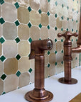 copper sprayer faucets against a tiled wall with green and beige tiles.