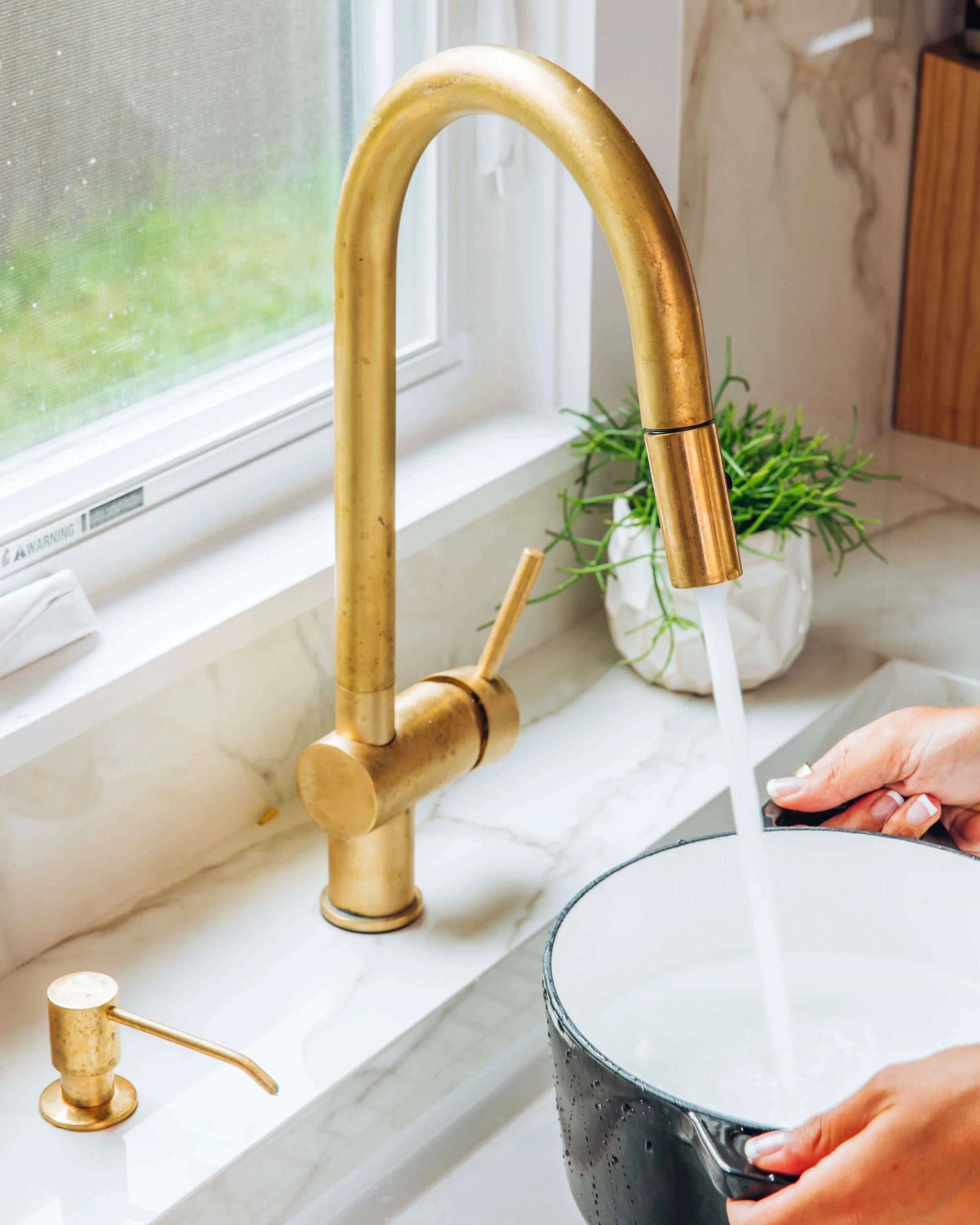 Brass kitchen faucet with running water into a black pot on a white countertop.