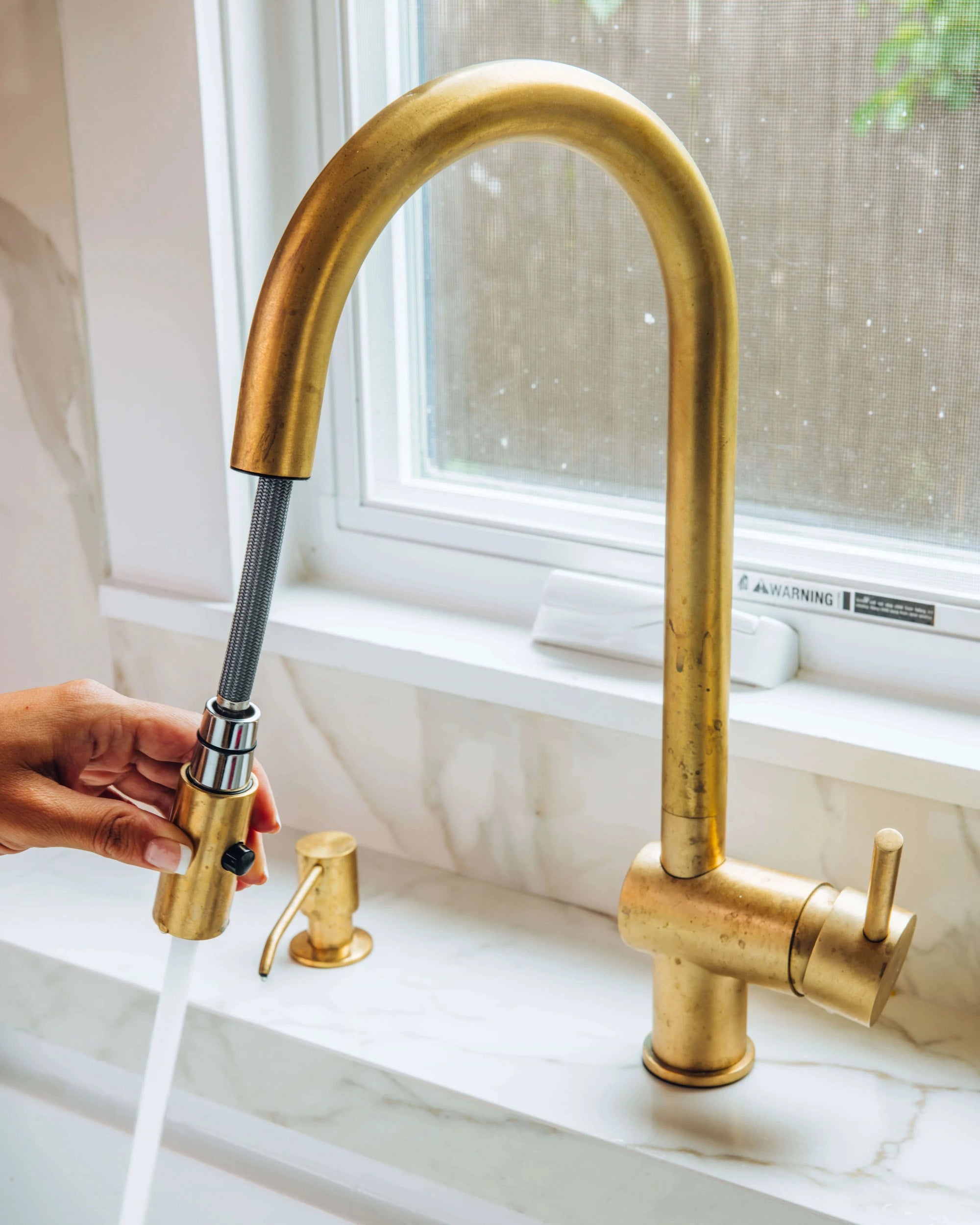 BRASS kitchen faucet with a hand turning on water, set against a neutral background.