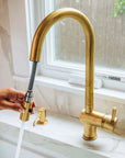 BRASS kitchen faucet with a hand turning on water, set against a neutral background.
