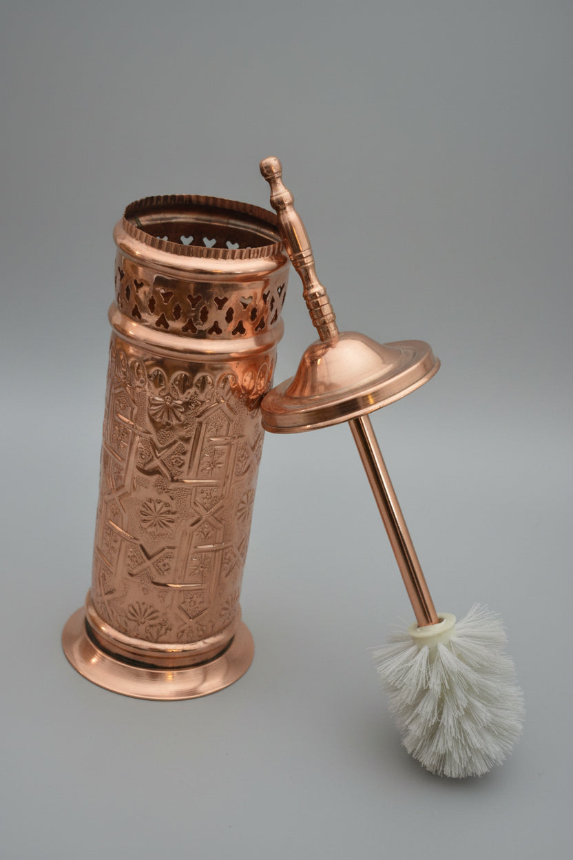 Copper toilet brush holder with intricate designs on a dark surface.