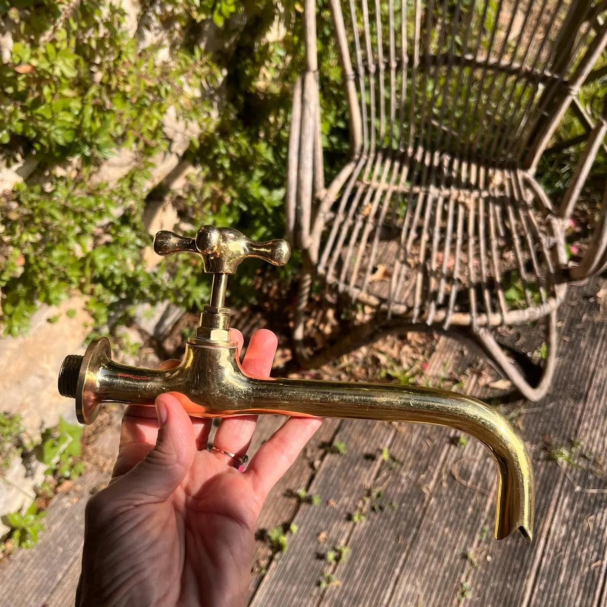 Person holding a vintage brass faucet outdoors with a wicker chair and greenery in the background.
