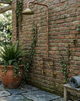 Outdoor shower system mounted on a brick wall with a potted plant and bench in the foreground.
