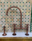 Bronze kitchen faucet with two handles against a mosaic tile backsplash.