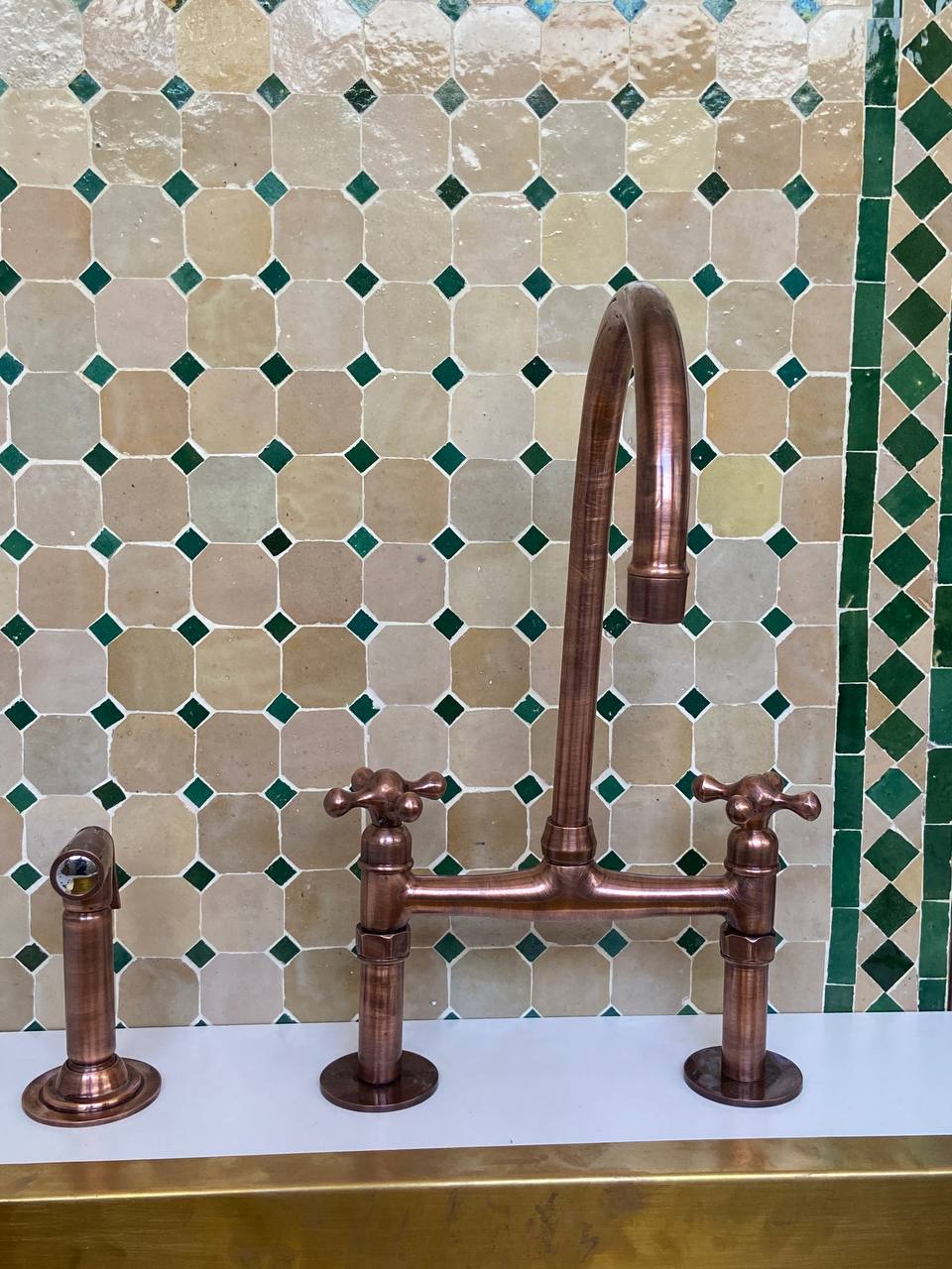 Copper kitchen faucet set against a mosaic tile wall with green and beige tiles.