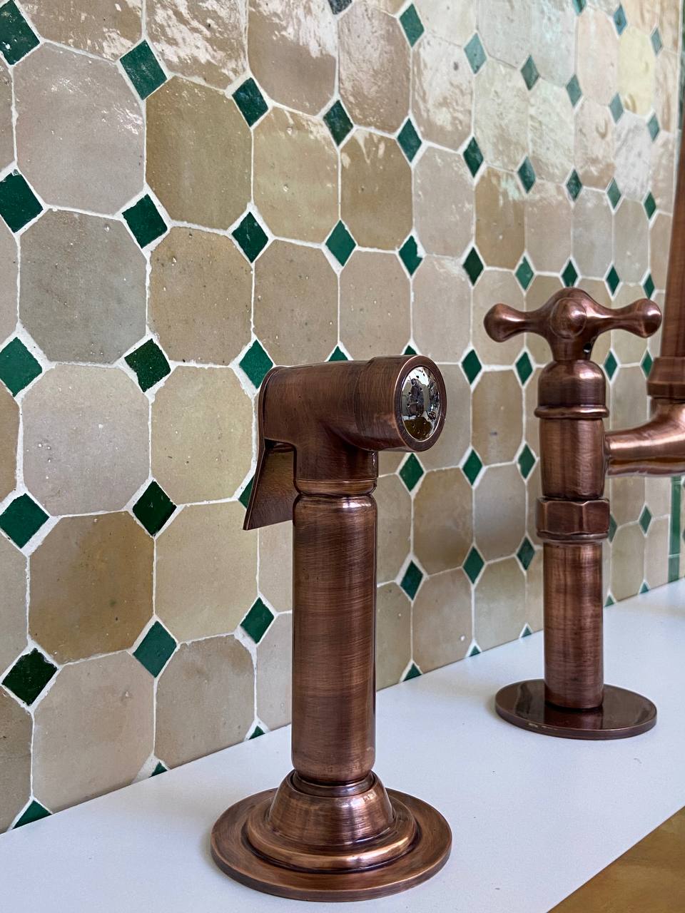 copper sprayer faucets against a tiled wall with green and beige tiles.