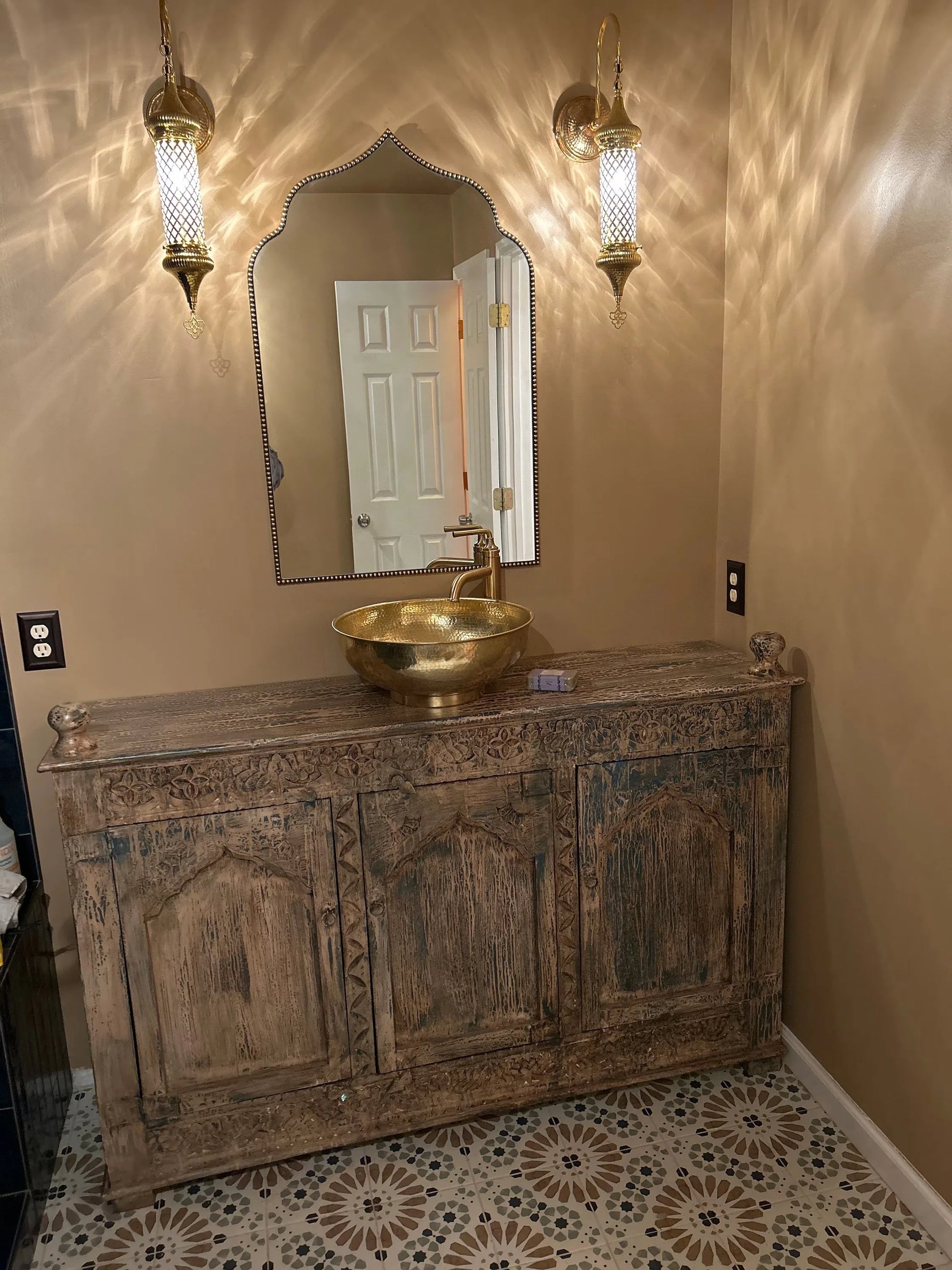 Bathroom with wooden vanity, gold sink, and decorative mirror.